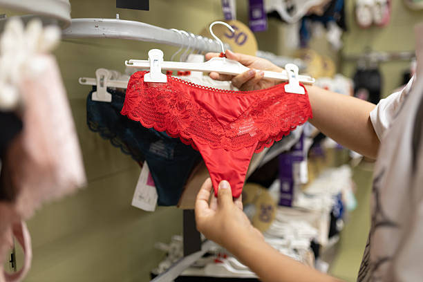 Un comprador sostiene una braga de encaje rojo brillante en una percha blanca y la examina entre otra ropa interior en un estante de la tienda. La escena captura moda minorista, compras y ropa íntima.