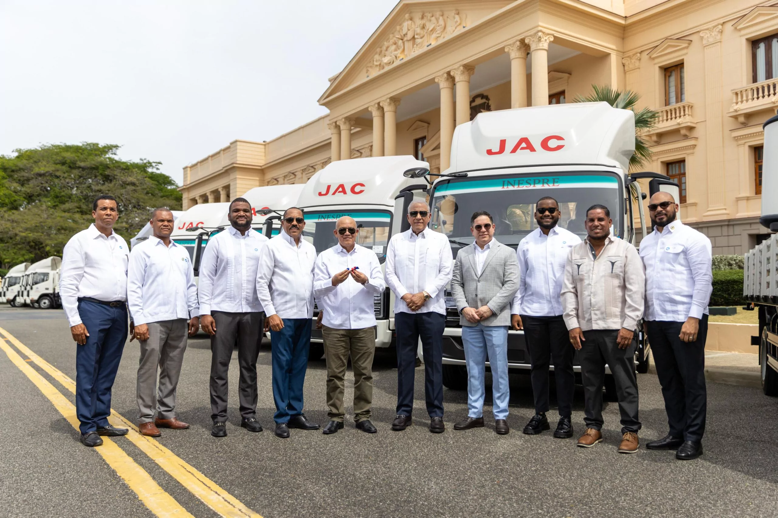 Representantes posan junto a camiones JAC frente a un edificio institucional en República Dominicana