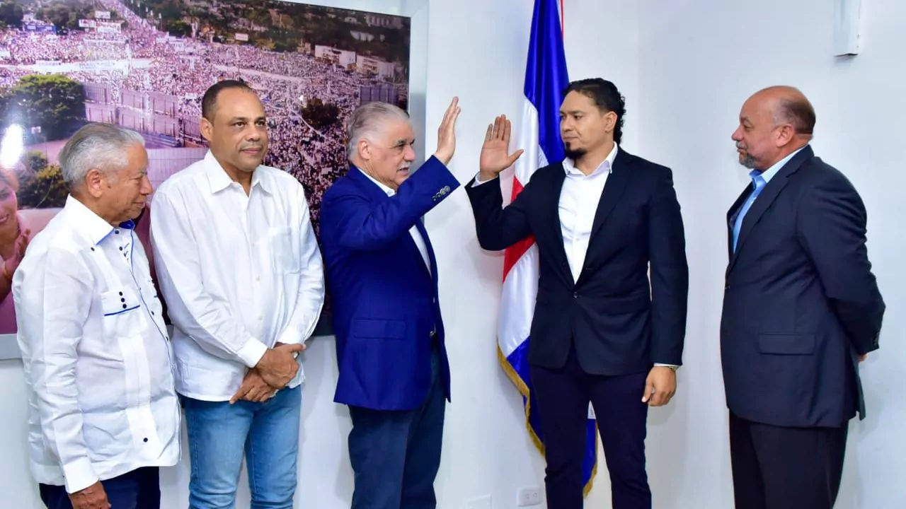 Hombres en reunión institucional junto a una bandera dominicana, uno saluda con la mano en alto