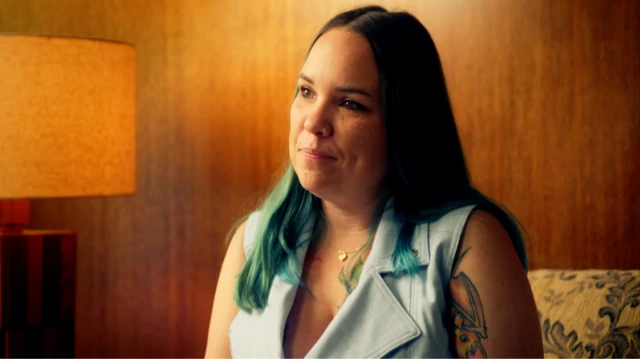 Taylor, una mujer de pelo largo y oscuro con un tatuaje que le cubre parte del brazo y la parte superior del pecho, lleva una blusa verde pálido sin mangas y está sentada en una habitación con paredes revestidas de madera y una lámpara naranja.