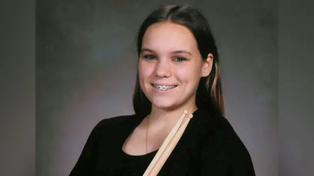 Taylor, a los 14 años, posa para un retrato. Viste una sencilla blusa negra de manga corta y sostiene dos baquetas de madera cruzadas sobre el pecho, mientras sorníe y en sus dientes se ven los aparatos que usaba en sus dientes.