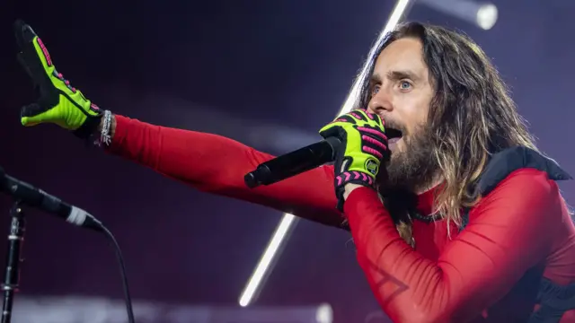 Jared Leto se encuentra bajo las luces del concierto, cantando con un micrófono de mano. Tiene el pelo largo y barba, y viste una camisa roja y ajustada, con guantes de color verde neón y rosa. Un brazo está extendido hacia el público, mientras que con el otro sostiene el micrófono cerca de su boca.  