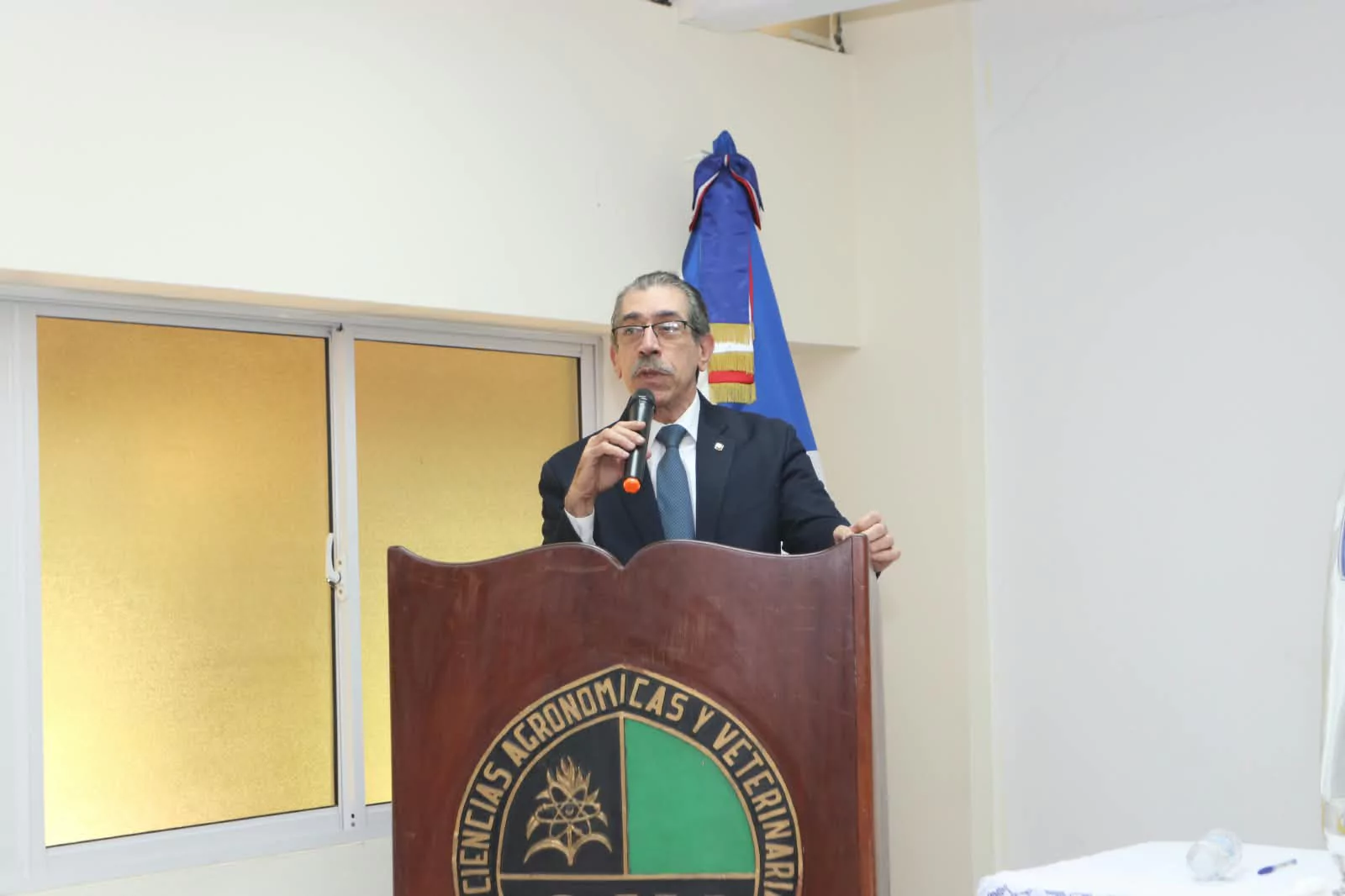 Hombre habla desde un podio en evento institucional con la bandera dominicana de fondo