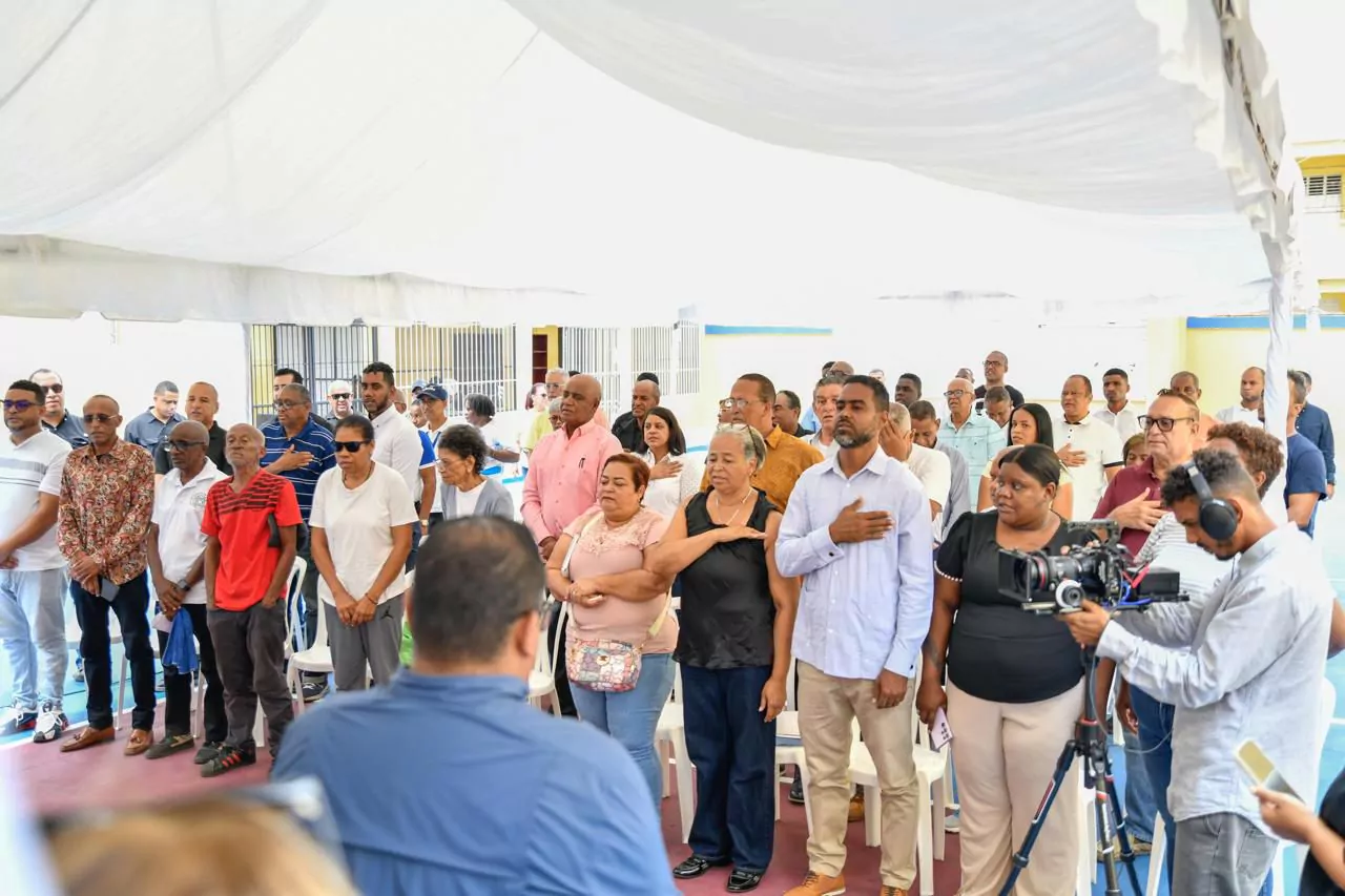 Los asistentes se encuentran durante un evento comunitario bajo una carpa, varios con las manos en el pecho, mientras un camarógrafo graba