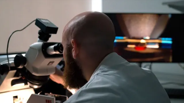 Una toma cinematográfica cercana desde detrás de un científico calvo y barbudo con una bata de laboratorio azul claro. Se inclina hacia adelante para mirar a través de un gran microscopio blanco, con el ocular negro pegado a su cara. En la parte superior hay un pequeño dispositivo parecido a una cámara, con un cable colgando hacia abajo. Una luz brillante ilumina la platina del microscopio y los hombros del hombre, mientras que el laboratorio circundante está a oscuras. Al fondo, un gran monitor muestra una imagen borrosa y ampliada en cálidos tonos marrones, atravesada por nítidas franjas horizontales de luz azul, blanca, naranja y roja.