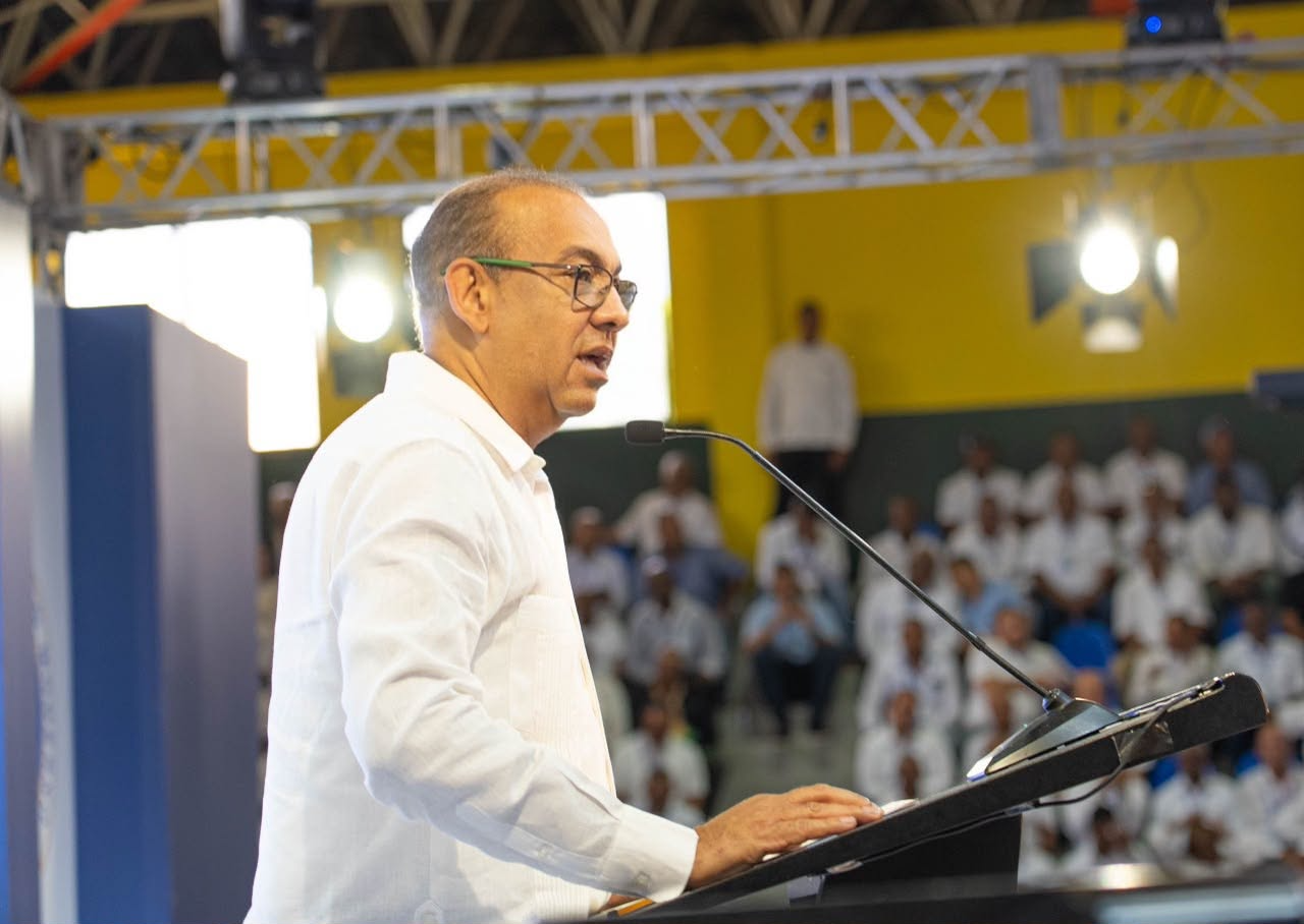 Un hombre con camisa blanca y gafas habla en un podio ante una audiencia en un evento público