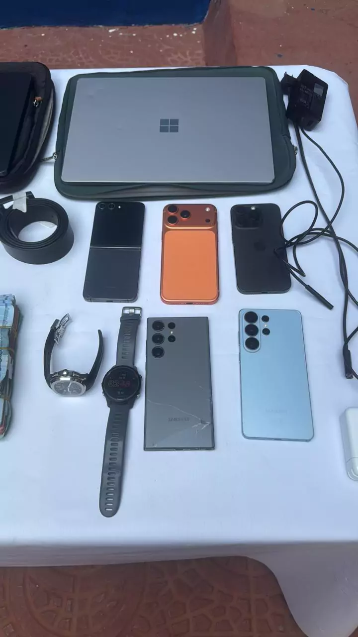 Computadora portátil, varios teléfonos celulares, relojes, cargadores, correa y dinero en efectivo colocados sobre una mesa blanca