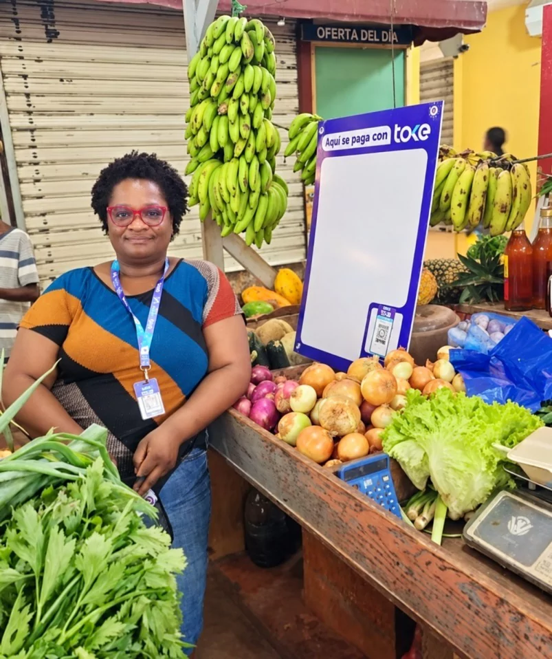 Vendedora en un puesto de frutas y verduras con un cartel de pago Toke, en el contexto del millón de usuarios de la plataforma