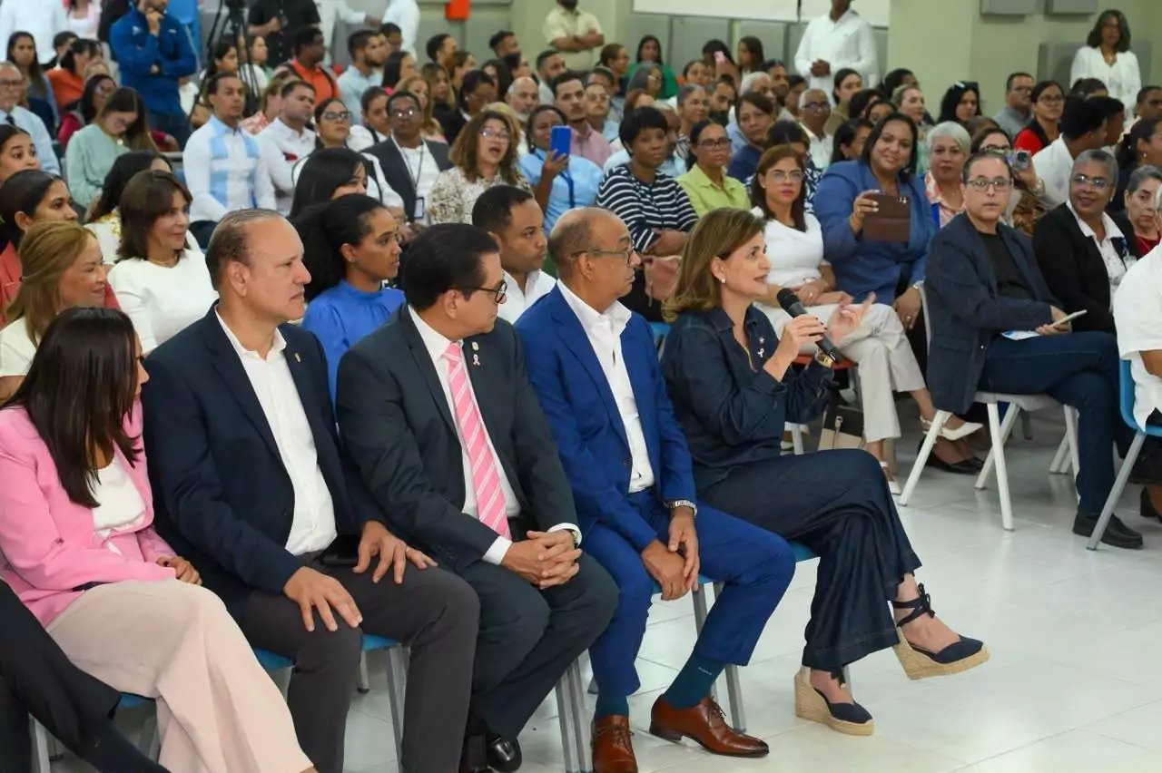 Funcionarios y asistentes en un acto público mientras una mujer interviene con un micrófono ante un numeroso público
